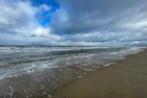 Strandzicht bij een vakantiehuis in Pandrup, Denemarken, met golven, nat zand en een bewolkte lucht.