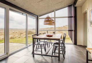 Salle à manger lumineuse avec grandes baies vitrées, vue sur les dunes et table pour six à Pandrup, Danemark.
