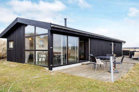 Zwart houten vakantiehuis in Pandrup, Denemarken, met grote ramen en terras met tuinmeubilair op gras.