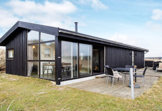 Schwarzes Ferienhaus in Pandrup, Dänemark, mit großen Fenstern und möblierter Terrasse auf Wiese.