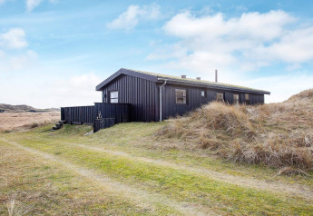 Casa vacanze nera per 6 persone a Pandrup, Danimarca, circondata da dune erbose sotto cielo azzurro.