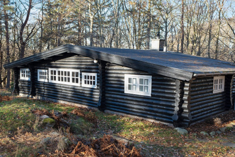 Cabaña negra de troncos con ventanas blancas rodeada de árboles en Frederiksværk, Dinamarca.