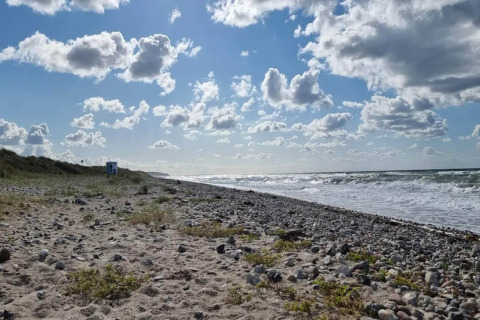 Rotsachtig en zanderig strand bij Frederiksværk, Denemarken, met blauwe lucht, wolken en zee.