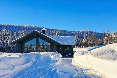 En hyggelig hytte dækket af sne i Passebekk, Sverige, omgivet af snedækkede skove og blå himmel.