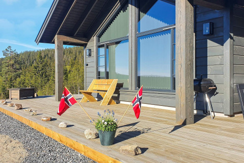 Wooden terrace of a modern chalet with Norwegian flags and grill, surrounded by lush green forest.