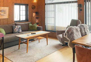Salon confortable dans un chalet suédois avec murs en bois, canapé, fauteuil et grandes fenêtres lumineuses.
