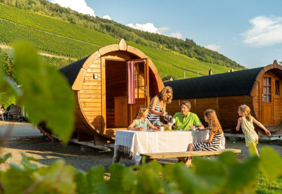 Familia comiendo frente a cabañas de barriles de vino en Camping Rissbach, rodeada de viñedos en Renania-Palatinado.