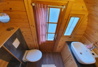 Petite salle de bain avec murs en bois, toilette, lavabo et porte avec rideau à carreaux en hébergement glamping.