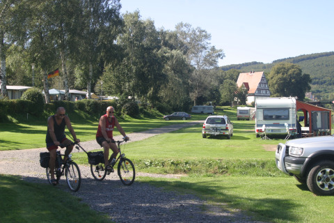 Dos hombres en bicicleta recorren Camping Gieselwerder con caravanas y césped verde en Hessen, Alemania.