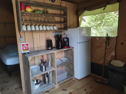 Area cucina nella tenda safari Domaine la Chabanne con mensole in legno, piatti, frigo e macchine da caffè.