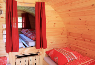 Innenansicht einer gemütlichen Holzhütte mit roten Vorhängen und Bettwäsche, Camping in Bayern.