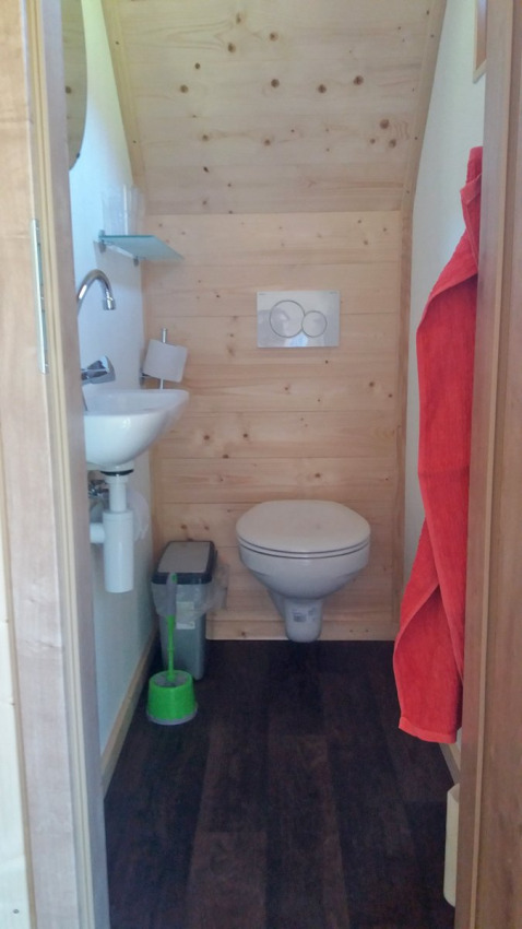 Baño pequeño con inodoro suspendido, lavabo, papelera y cepillo verde en lodge de glamping de madera.