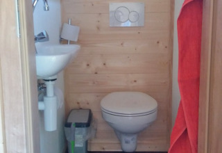 Small bathroom with wall-hung toilet, sink, waste bin, and green toilet brush in a wood glamping lodge.