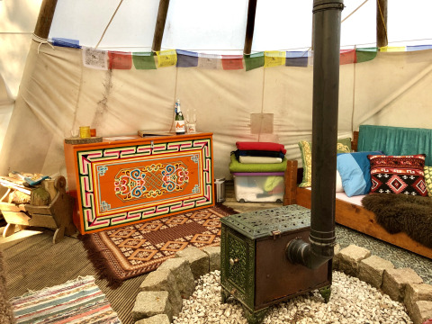 Indretningen af en hyggelig tipi med brændeovn, farverige tæpper og puder, i glampingstil.