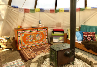 Gemütliches Interieur eines Tipis mit Holzofen, bunten Kissen und Teppichen im Glamping-Stil.