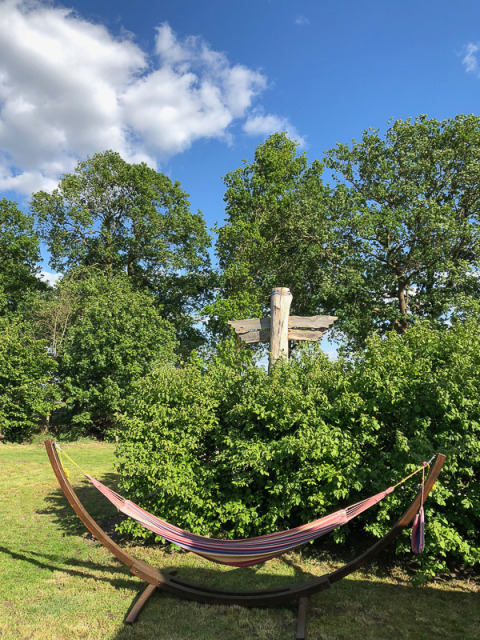 Outdoor glamping area with a colorful hammock, green shrubs, signpost, and trees under a blue sky with clouds.