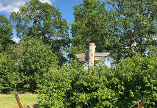 Area glamping all’aperto con amaca colorata, alberi, cespugli verdi e segnale sotto cielo blu e nuvole.