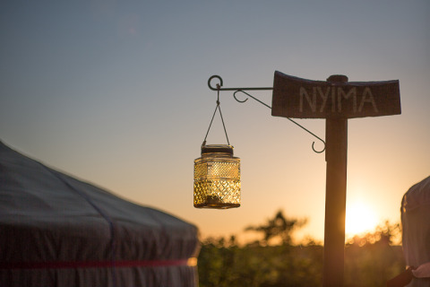 Coucher de soleil sur un site de glamping, pancarte 'NYIMA' et lanterne suspendue devant une yourte.