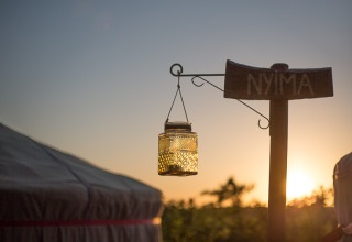 Solnedgang ved et glampingsted med et skilt, hvor der står 'NYIMA', og en hængende lanterne udenfor en yurt.