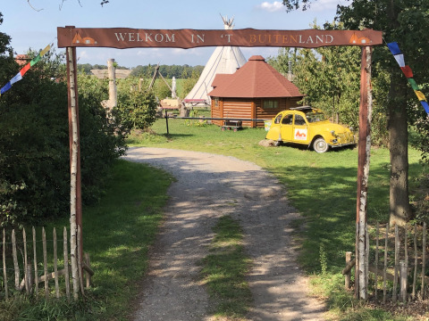 Indgang til glampingplads med tipi, træhytte og gul bil under skiltet 'Welkom in 't Buitenland'.