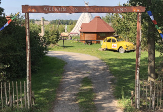 Indgang til glampingplads med tipi, træhytte og gul bil under skiltet 'Welkom in 't Buitenland'.