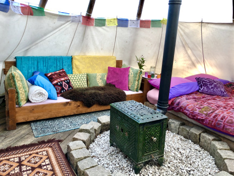 Colorful glamping tipi interior with sofa, bed, wood stove, many pillows, and decorative banners above.