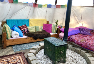 Colorful glamping tipi interior with sofa, bed, wood stove, many pillows, and decorative banners above.