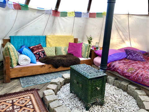 Kleurrijk glamping tipi-interieur met bank, bed, kachel, veel kussens en decoratieve vlaggetjes erboven.