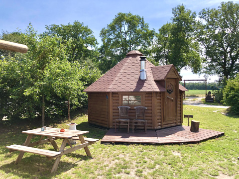 Glamping accommodation in nature with a wooden cabin, picnic table, and chairs under blue sky and trees.