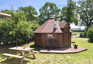 Glamping accommodation in nature with a wooden cabin, picnic table, and chairs under blue sky and trees.
