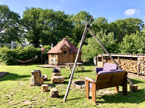Zona de glamping acogedora con Kota finlandesa, fogata, troncos, banco, leña apilada y hamaca en la naturaleza.