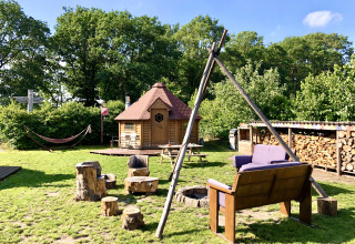 Knusse glampingplek met Finse kota, vuurplaats, boomstamstoelen, bank, houtopslag en hangmat in de natuur.