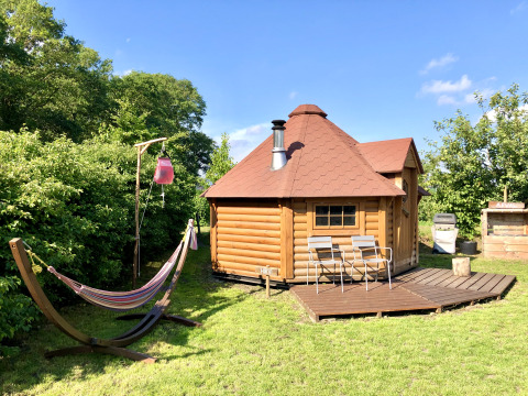 Glamping-Unterkunft: Finnische Kota mit Terrasse, Hängematte und Gartenstühlen bei Sonnenschein.