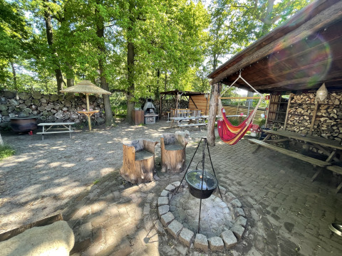 Gemütlicher Glampingplatz im Freien mit Lagerfeuer, Hängematte, Holzmöbeln und gestapeltem Brennholz.