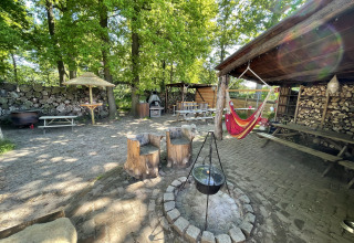 Outdoor glamping area with hammock, fire pit, wooden chairs and tables, surrounded by lush green trees.