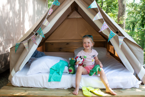 Petite fille assise sur un lit dans une tente safari du Chianti Glamping Resort, souriante avec des peluches.