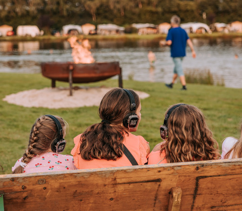 Kinder mit Kopfhörern sitzen auf einer Bank am Lagerfeuer am Wasser bei Recreatiepark de Achterste Hoef Glamping.