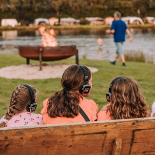 Children with headphones sit on a bench by a firepit at the lake at Recreatiepark de Achterste Hoef glamping.