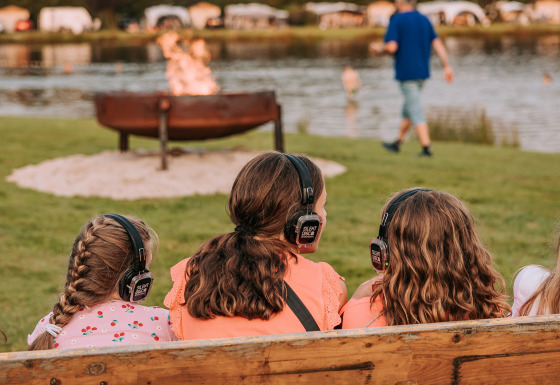 Children with headphones sit on a bench by a firepit at the lake at Recreatiepark de Achterste Hoef glamping.