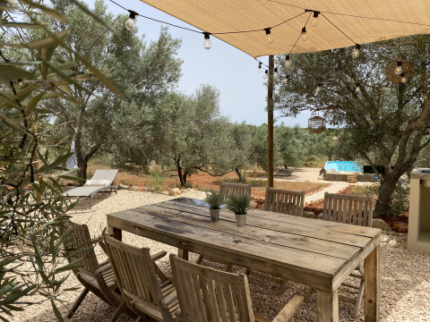Mesa de comedor al aire libre bajo toldo, luces, sillas de madera, olivares y piscina en Algarve Olive Tree Lodge.