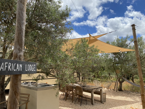 Udendørs glampingområde ved Algarve Olive Tree Lodge med træmøbler, solsejl og skiltet 'African Lodge'.