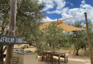Outdoor glamping area at Algarve Olive Tree Lodge with wooden furniture, shade sails and 'African Lodge' sign.
