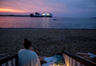 Person sidder i strandkurv ved Kieler Fjord under solnedgang, med skib på vandet og glampingstemning.