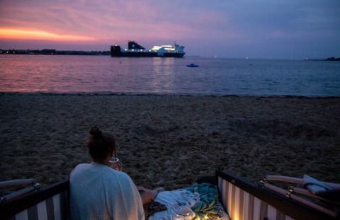 Persoon zit in een strandstoel aan Kieler Fjord bij zonsondergang, schip op het water, glamping sfeer.