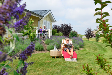 Man rust op ligstoel terwijl familie geniet bij chalet op Camping en Vakantiepark Brilon, Noordrijn-Westfalen.