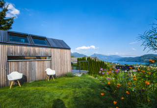 Wooden glamping accommodation with chairs, flower garden, and mountain-lake view in Austria.