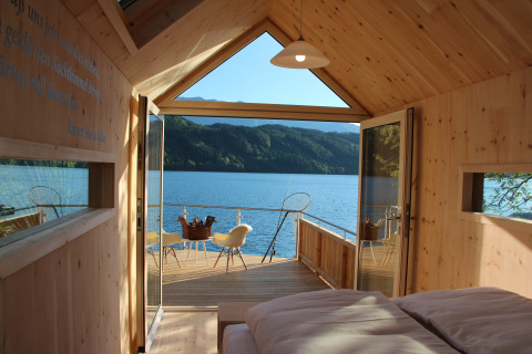 Cabaña glamping de lujo con interior de madera, puertas abiertas a terraza junto al lago y vista a montañas.