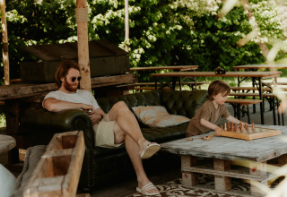 Een man ontspant op een bank, terwijl een kind buiten schaakt bij glamping REBL Outdoor in Friesland.