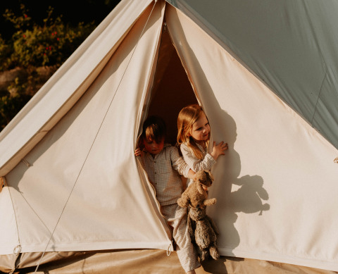 Due bambini sbirciano fuori da una tenda glamping presso REBL Outdoor – Bell tenten Friesland al sole del mattino.