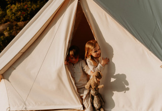 Due bambini sbirciano fuori da una tenda glamping presso REBL Outdoor – Bell tenten Friesland al sole del mattino.